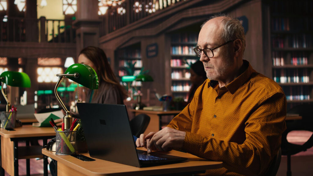 Senior man in a yellow shirt working on a laptop in a large, dimly lit library with green desk lamps, researching international patent application requirements.
