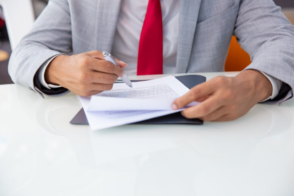 A businessman in a grey suit and red tie signing a legal contract to sell or license a registered industrial design.