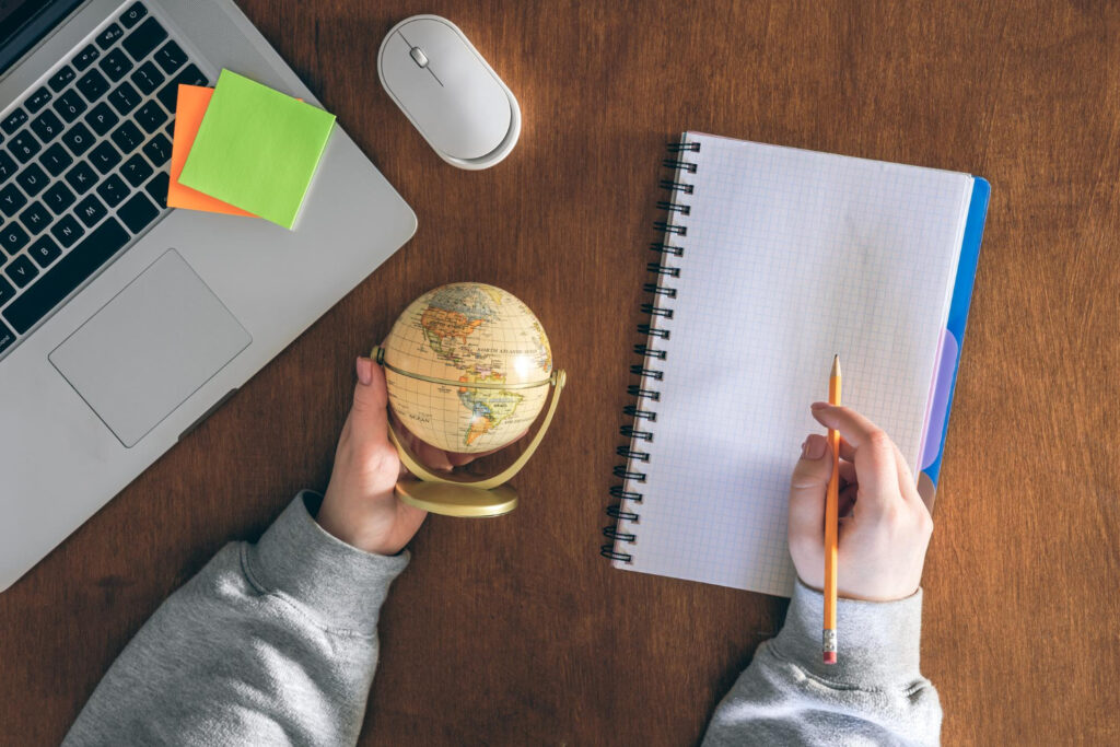 Hands holding a small globe next to a laptop, mouse, and notebook on a wooden desk, illustrating international trademark filing and global business protection.
